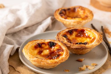 Geleneksel Portekiz usulü yumurta muhallebi hamuru olan üç altın-kahverengi Pastel de Nata, açık gri bir tabakta, yanında küçük bir tarçın ve vanilya çekirdeği ile birlikte sergilenmektedir..