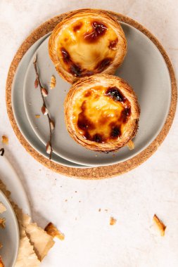 İki Portekiz muhallebi tartı, pastel de nata, gri bir tabakta karamelize edilmiş, hafif bir yüzeyde bir mantar paspasın üzerine yerleştirilmiş, tomurcuklu küçük bir dalla birlikte. Üst görünüm