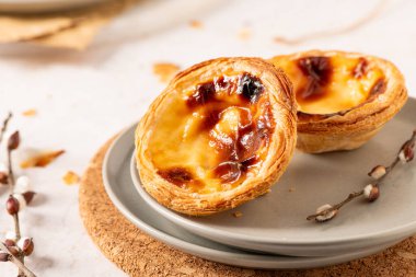 İki Portekizli muhallebi tartı, pastel de nata, gri bir tabakta karamelize edilmiş, hafif bir yüzeyde bir mantar paspasın üzerine yerleştirilmiş, tomurcuklu küçük bir dal eşliğinde.