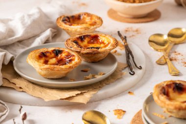 Portekiz kremalı tart koleksiyonu olan Pastel de Nata, yanında tarçın tozu, vanilya çekirdeği ve küçük altın kaşıklarla birlikte açık gri tabaklarda sergilenmektedir. Söğüt dalları bir bahar dokunuşu ekler.