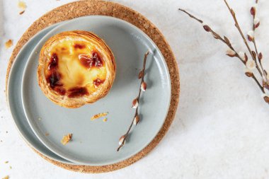 Bir Pastel de Nata, Portekiz usulü yumurta kremalı tart, gri bir tabakta narin söğüt dallarıyla oturur. Sahne, arka planda hafif bir mantar altlığı üzerine kurulmuş. Üst görünüm