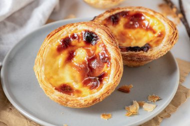 İki Portekizli muhallebi tartı, pastel de nata, gri bir tabakta karamelize edilmiş, hafif bir yüzeyde bir mantar paspasın üzerine yerleştirilmiş, tomurcuklu küçük bir dal eşliğinde.