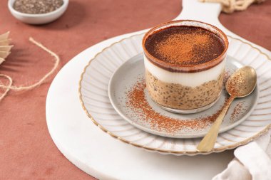 Tiramisu chia pudingi beyaz bir tabakta, beyaz bir tabakta, kakao tozuyla süslenmiş, kahverengi bir yüzeye kurulmuş, arka planda elek ve kakao tozu var.