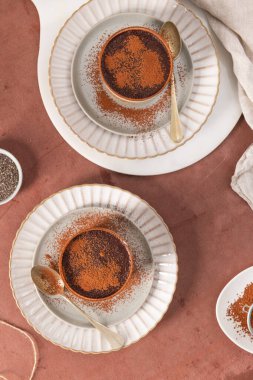 Beyaz tabaklara kakao tozu serpiştirilmiş iki çikolatalı tatlı, altın kaşıklarla birlikte, kahverengi bir yüzeye kurulmuş, arka planda beyaz bir bez parçası var. Üst görünüm