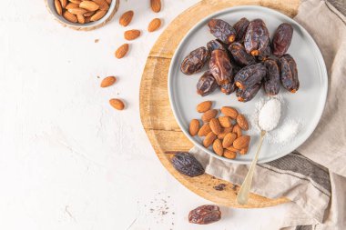 Medjool dates and almonds on a gray plate with a spoonful of shredded coconut, placed on a wooden board with a beige cloth in the background. Top view clipart