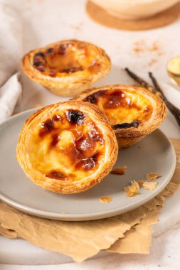 Geleneksel Portekiz usulü yumurta muhallebi hamuru olan üç altın-kahverengi Pastel de Nata, açık gri bir tabakta, yanında küçük bir tarçın ve vanilya çekirdeği ile birlikte sergilenmektedir..