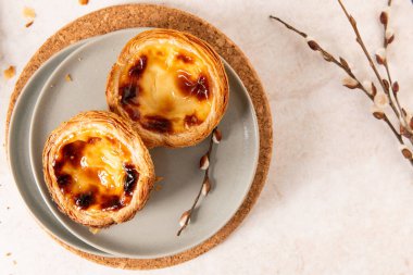 İki Portekiz muhallebi tartı, pastel de nata, gri bir tabakta karamelize edilmiş, hafif bir yüzeyde bir mantar paspasın üzerine yerleştirilmiş, tomurcuklu küçük bir dalla birlikte. Üst görünüm