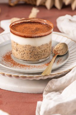 Tiramisu chia pudingi beyaz bir tabakta, beyaz bir tabakta, kakao tozuyla süslenmiş, kahverengi bir yüzeye kurulmuş, arka planda elek ve kakao tozu var.