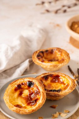 Geleneksel Portekiz usulü yumurta muhallebi hamuru olan üç altın-kahverengi Pastel de Nata, açık gri bir tabakta, yanında küçük bir tarçın ve vanilya çekirdeği ile birlikte sergilenmektedir..