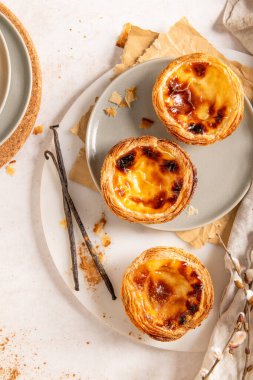 Üç Portekiz kremalı turtası, Pastel de Natas, açık arkaplanda vanilyalı fasulye ve söğüt dallarıyla birlikte gri bir tabakta yer almaktadır. Üst görünüm