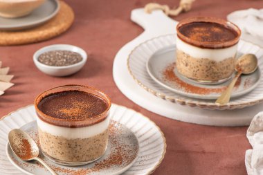 Üstü kremalı yoğurt ve kakao tozuyla kaplı Chia pudingi. Kırsal bir masada, zarif tabaklarda servis edilen cam kavanozlarda servis ediliyor.