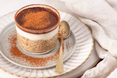 Bardaktaki tiramisu chia pudingi, kakao tozuyla kaplı, beyaz bir tabakta altın bir kaşık ve yanında bej bir peçete.