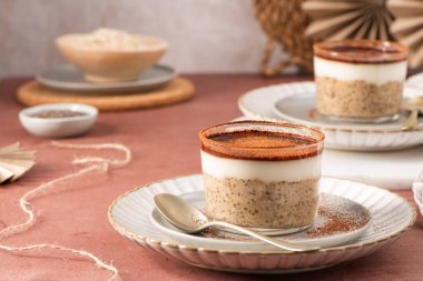 Kremalı tiramisu gece yulafı ve yoğurt ve kakao tozuyla kaplanmış chia pudingi, beyaz kaşıklarla servis edilen zarif cam bardaklarda, kırsal bir masada.