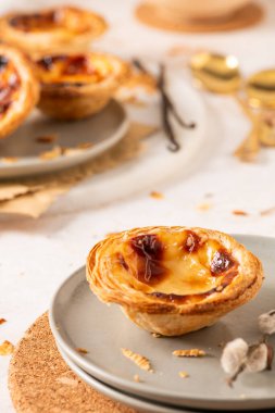 Portekiz kremalı turtası, Pastel de Nata, bir mantar altlığının üzerinde duran açık gri bir tabağa odaklanmıştır. Diğer tartlar, vanilyalı fasulyeler ve söğüt çiçekleri yumuşak arka planda görülebilir..