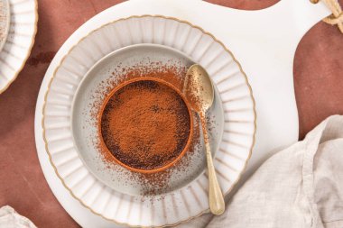 Beyaz bir tabakta kakao tozu tozlu kakao tatlısı yanında da kahverengi bir yüzey ve etrafa saçılmış kakao. Üst görünüm