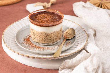Üzerinde kakao tozu olan tiramisu chia pudingi, beyaz bir tabakta cam bir kavanozda altın kaşıkla servis ediliyor, ve yakınlarda küçük bir kase chia çekirdeği var.