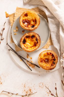 Üç Portekiz kremalı turtası, Pastel de Natas, açık arkaplanda vanilyalı fasulye ve söğüt dallarıyla birlikte gri bir tabakta yer almaktadır. Üst görünüm