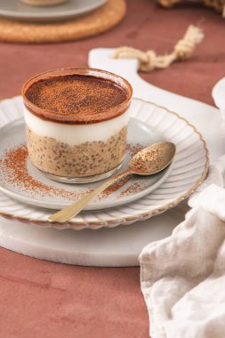 Tiramisu chia pudingi beyaz bir tabakta, beyaz bir tabakta, kakao tozuyla süslenmiş, kahverengi bir yüzeye kurulmuş, arka planda elek ve kakao tozu var.