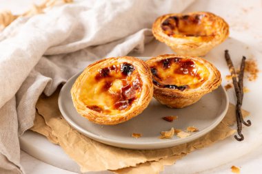 Geleneksel Portekiz usulü yumurta muhallebi hamuru olan üç altın-kahverengi Pastel de Nata, açık gri bir tabakta, yanında küçük bir tarçın ve vanilya çekirdeği ile birlikte sergilenmektedir..