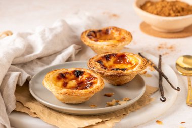 Geleneksel Portekiz usulü yumurta muhallebi hamuru olan üç altın-kahverengi Pastel de Nata, açık gri bir tabakta, yanında küçük bir tarçın ve vanilya çekirdeği ile birlikte sergilenmektedir..