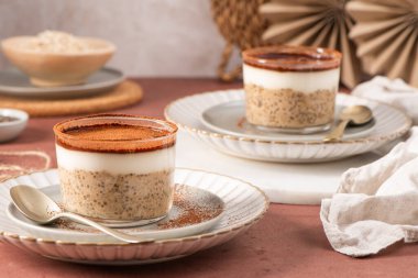 Kremalı tiramisu gece yulafı ve yoğurt ve kakao tozuyla kaplanmış chia pudingi, beyaz kaşıklarla servis edilen zarif cam bardaklarda, kırsal bir masada.