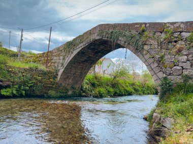 Travassos Ortaçağ Köprüsü. Tek kemerli eski bir taş köprü, Portekiz 'in Felgueiras kentindeki Jugueiros şehrinde bulutlu bir gökyüzünün altında yemyeşil bitki örtüsüyle çevrili temiz ve sığ bir nehir boyunca uzanır.