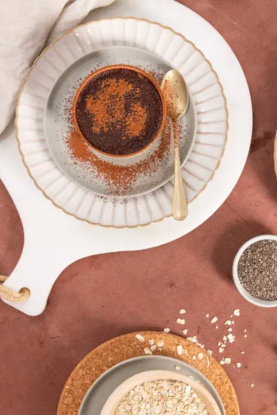 Beyaz bir tabakta kakao tozu ile çikolata tatlısı, yanında altın bir kaşık, bir kasede yulaf, küçük bir tabakta chia tohumu ve beyaz bir bez. Üst görünüm