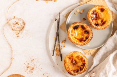 Üç Portekiz kremalı turtası, Pastel de Natas, açık arkaplanda vanilyalı fasulye ve söğüt dallarıyla birlikte gri bir tabakta yer almaktadır. Üst görünüm