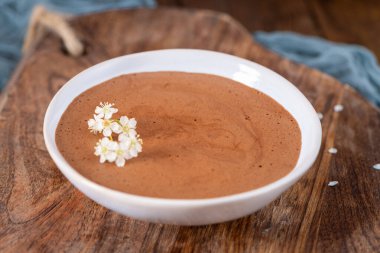 Beyaz bir kasede yumuşak çikolatalı mus, narin beyaz çiçeklerle süslenmiş, yumuşak mavi kumaşlı bir ahşap tahta üzerinde sunuluyor.