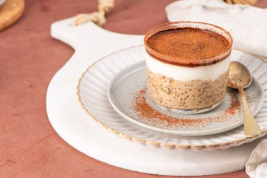 Üzerinde kakao tozu olan tiramisu chia pudingi, beyaz bir tabakta cam bir kavanozda altın kaşıkla servis ediliyor, ve yakınlarda küçük bir kase chia çekirdeği var.