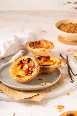 Geleneksel Portekiz usulü yumurta muhallebi hamuru olan üç altın-kahverengi Pastel de Nata, açık gri bir tabakta, yanında küçük bir tarçın ve vanilya çekirdeği ile birlikte sergilenmektedir..