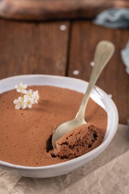 Beyaz bir kasede ipek çikolatalı mus, küçük beyaz çiçeklerle süslenmiş bir kaşık kaldırılmış, buruşuk parşömen kağıdı ve kırsal ahşap bir masa üzerinde sunulmuş.