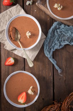 Üç beyaz kase yumuşak çikolatalı mus, biri altın kaşıkla, ahşap bir masada taze çilekler ve narin beyaz çiçeklerle güzel bir şekilde düzenlenmiştir.