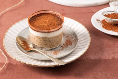 Tiramisu chia pudingi beyaz bir tabakta, beyaz bir tabakta, kakao tozuyla süslenmiş, kahverengi bir yüzeye kurulmuş, arka planda elek ve kakao tozu var.