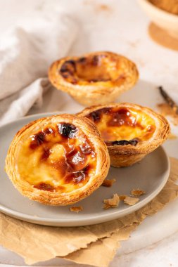 Geleneksel Portekiz usulü yumurta muhallebi hamuru olan üç altın-kahverengi Pastel de Nata, açık gri bir tabakta, yanında küçük bir tarçın ve vanilya çekirdeği ile birlikte sergilenmektedir..