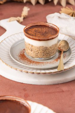 Tiramisu chia pudingi beyaz bir tabakta, beyaz bir tabakta, kakao tozuyla süslenmiş, kahverengi bir yüzeye kurulmuş, arka planda elek ve kakao tozu var.
