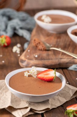 Taze çilek ve küçük beyaz çiçeklerle süslenmiş pürüzsüz çikolatalı mus tabakları kırsal ahşap bir tahta ve parşömen kağıdına dizilmiştir.