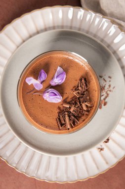 Çok yakın bir tepeden çekimde çikolata parçacıkları ve dekoratif tabaklar üzerinde mor çiçeklerle süslenmiş kremalı çikolatalı kremayı bir bardakta yakalıyor..