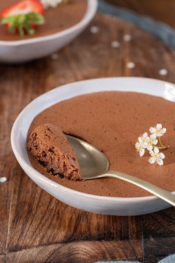 Beyaz bir kasede kremalı çikolatalı mus, bir ısırık alan altın bir kaşık, narin beyaz çiçeklerle süslenmiş, sıcak ahşap bir yüzeyde dinleniyor.