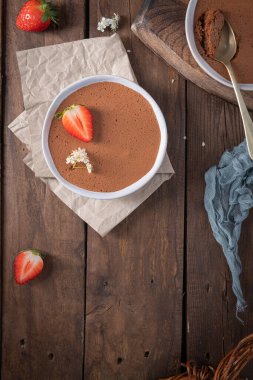 Taze çilek ve narin beyaz çiçeklerle bezenmiş beyaz bir kasede yumuşak bir çikolatalı mus, altın kaşıkla kırsal bir ahşap masada duruyor.