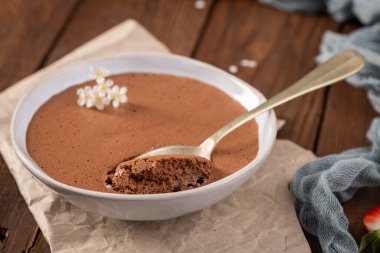 Sığ beyaz bir tabakta zarif çikolatalı mus. Isırığı kepçeleyen altın bir kaşık. Küçük beyaz çiçeklerle süslenmiş.