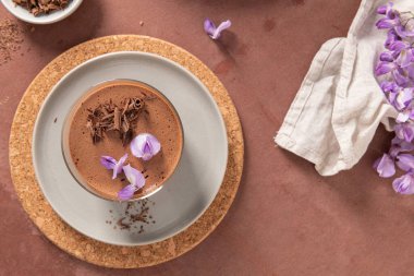 Genel bir bakış açısında bir bardakta kremalı çikolatalı mus, mantar kaplamalı bir tabak, bir kase talaş ve mor çiçekler bulunur..