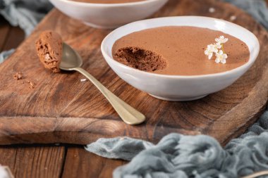 Beyaz kaselerde zengin çikolatalı mus tabakları, bir kaşık dolusu kaldırılmış, narin beyaz çiçeklerle süslenmiş, yumuşak mavi kumaşlı kırsal ahşap bir tahtanın üzerinde sunulmuş.