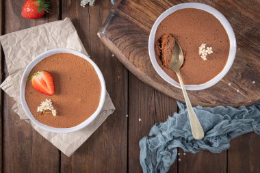 İki kase yumuşak çikolatalı mus, biri çilekle süslenmiş, ahşap bir yüzey üzerinde altın bir kaşık ve narin çiçeklerle ustalıkla düzenlenir.