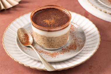 Üzerinde kakao tozu olan tiramisu chia pudingi, beyaz bir tabakta cam bir kavanozda altın kaşıkla servis ediliyor, ve yakınlarda küçük bir kase chia çekirdeği var.