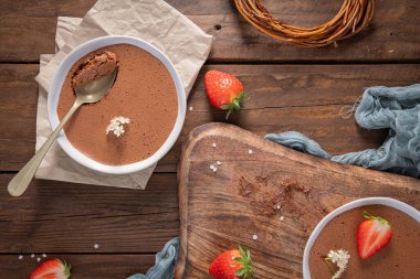 İki beyaz kase kremalı çikolatalı mus, biri altın kaşıklı, taze çilekler ve narin beyaz çiçeklerle kırsal ahşap bir masada sunuluyor.