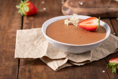 Beyaz bir kasedeki zarif çikolata mus, taze çilek dilimi ve ince beyaz çiçeklerle süslenmiş, sıcak ahşap bir yüzey üzerinde dokulu parşömen kağıdına yaslanmış.