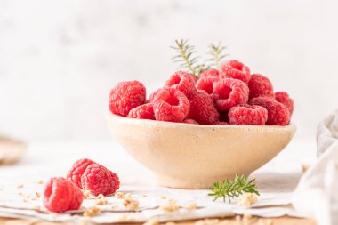 Dolgun, taze ahududu dolu bir kırsal seramik kase dağınık tahıl gevreğiyle çevrili beyaz bir masada oturur.