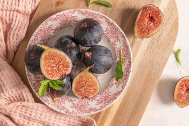 Bir tabak taze incir, ahşap bir tahta, taze stevia yaprakları ve yumuşak ışıkta pembe bir peçete.