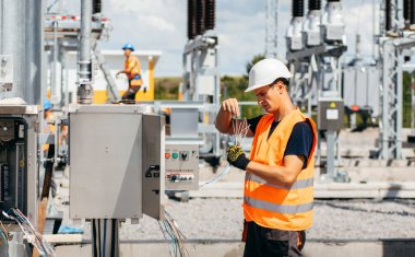 Koruyucu kasklı elektrikçi yüksek voltajlı elektrik hatları üzerinde çalışıyor. Elektrik şebekesine hizmet eden çok yetenekli işçiler. Güç kuleleri olan modern enerji santrali